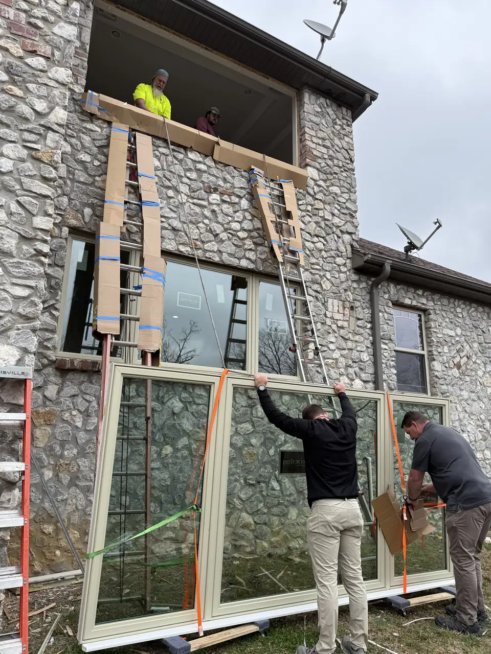 Crew lifting a large window unit into place on a two-story stone home